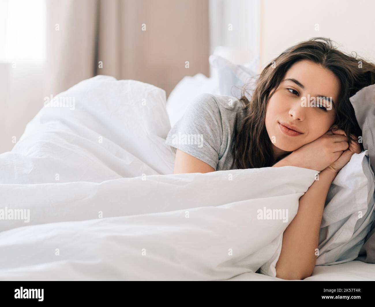 Cozy morning. Sunday relaxation. Home rest. Peaceful thoughtful daydreaming brunette woman enjoying lying in bed alone with white blanket in light bed Stock Photo
