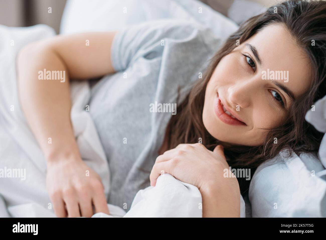 Good morning. Weekend rest. Home relaxation. Beauty health. Happy relaxed lazy woman enjoying lying awake in bed smiling looking at camera in light be Stock Photo