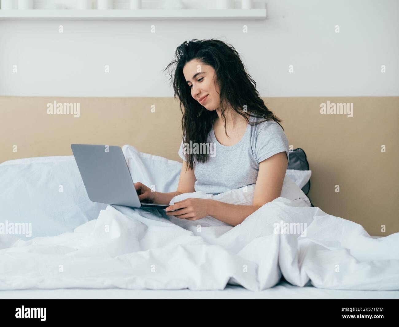 Morning chat. Online communication. Modern technology. Home leisure. Relaxed happy woman enjoying reading message using laptop in bed in light bedroom Stock Photo