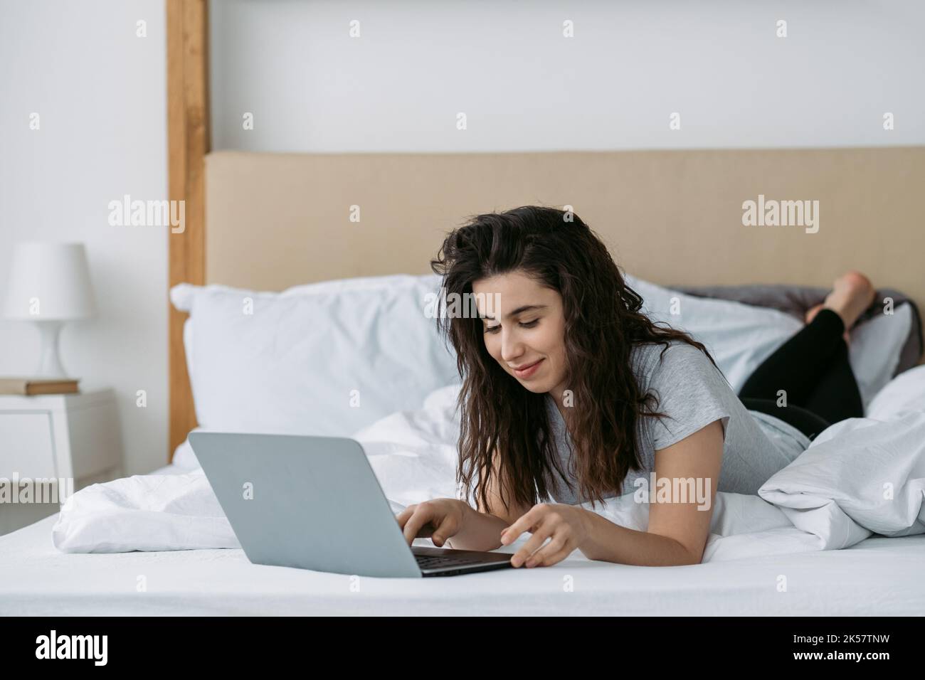 Morning chat. Virtual communication. Social media. Digital weekend. Relaxed woman enjoying speaking online using laptop network lying on bed in light Stock Photo