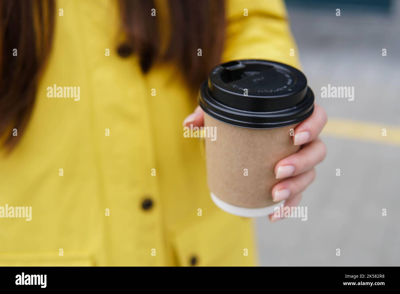 Close up of young woman in yellow raincoat holding paper cup of coffee takeaway in the street. Stock Photo