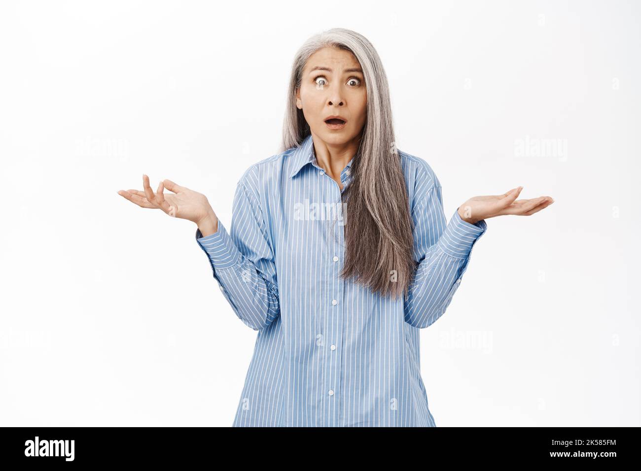 Shocked and confused senior asian woman, middle aged korean lady shrugging shoulders and looking puzzled, standing over white background Stock Photo