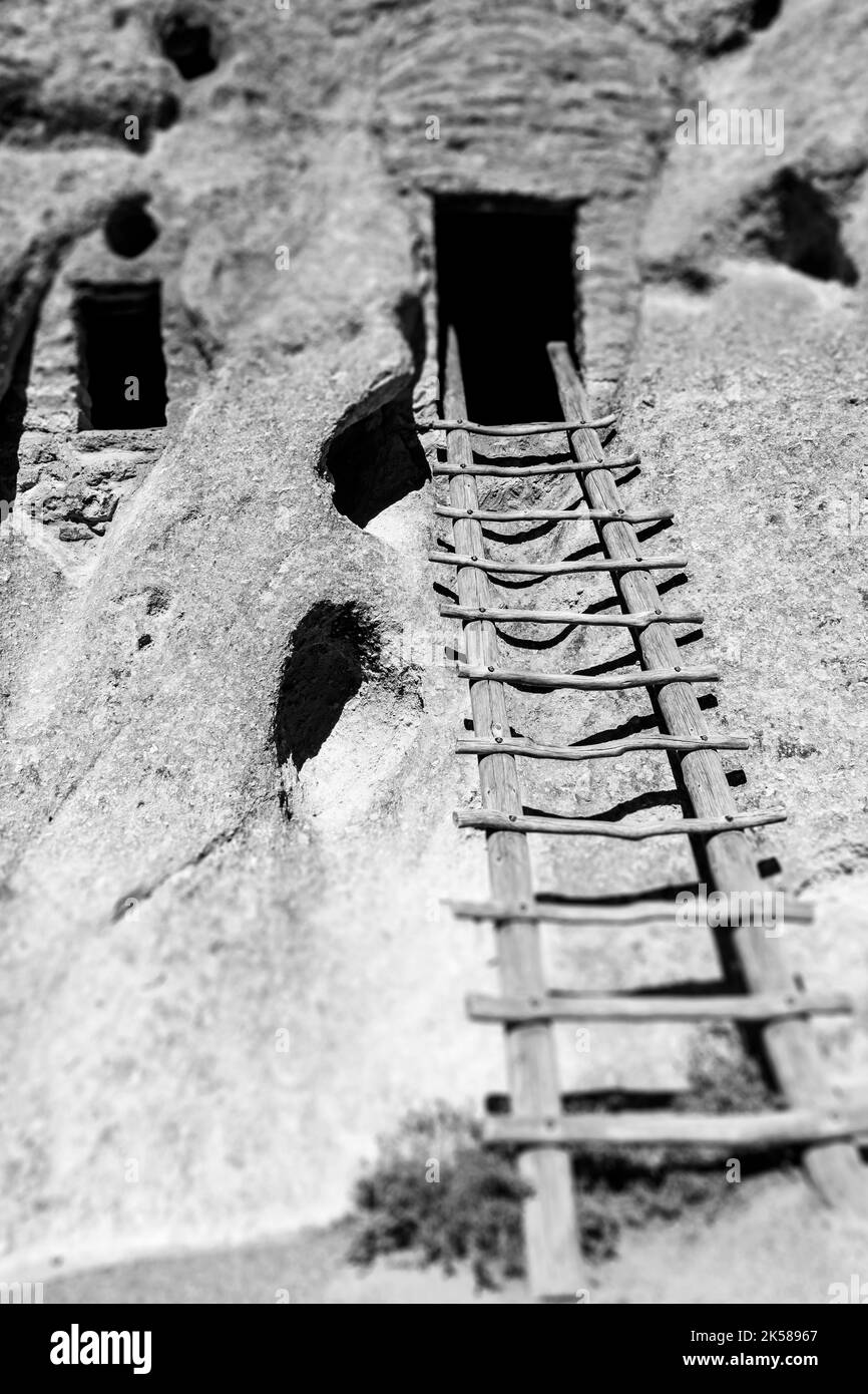A black and white image of a wooden ladder leads to the open door of a cave dwelling in Bandelier National Monument, New Mexico, USA. Stock Photo