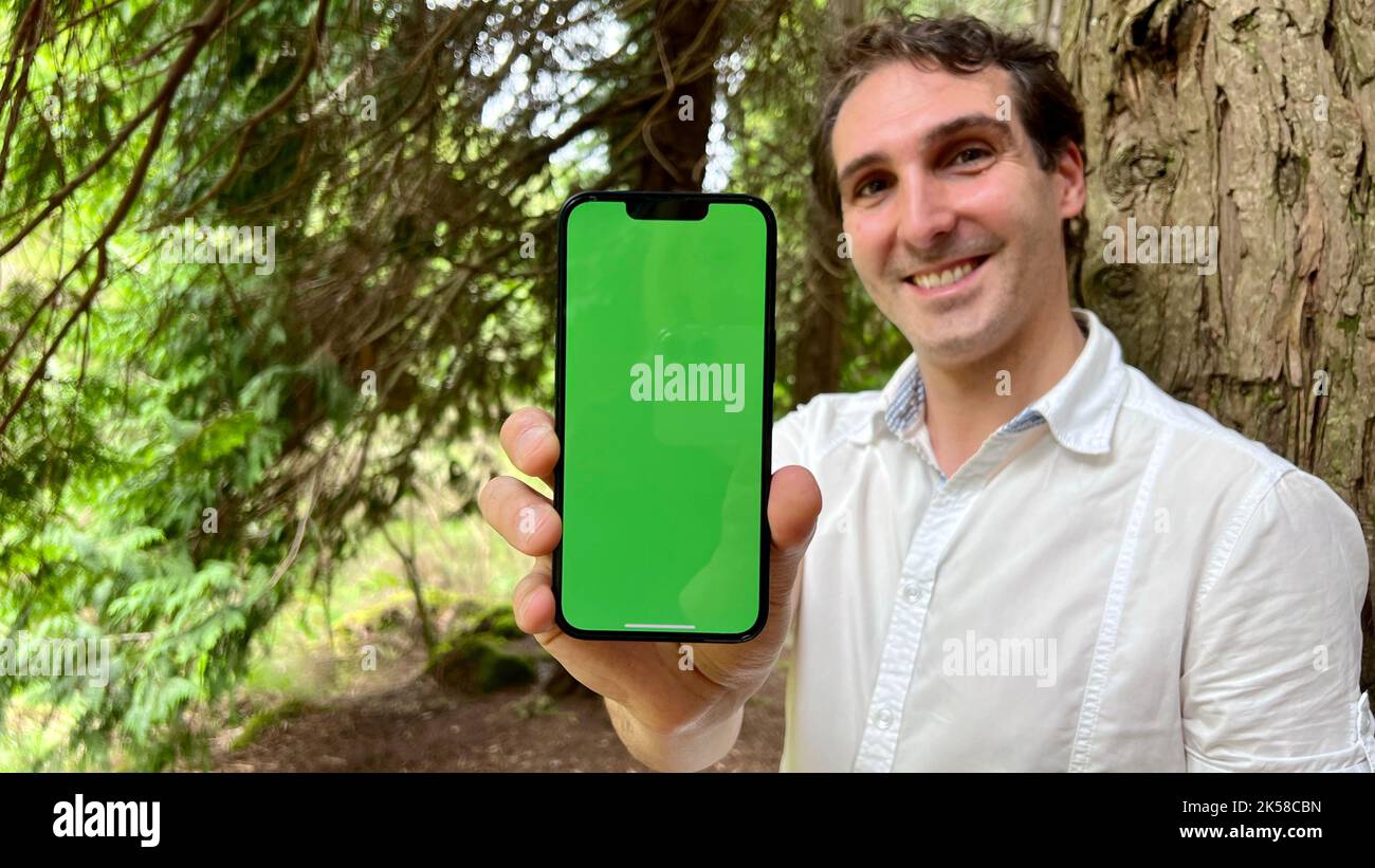 very handsome man french italy not holding a phone with a green tap standing in the woods in a white shirt and smiling showing a finger on the screen can use for advertising. High quality 4k footage Stock Photo