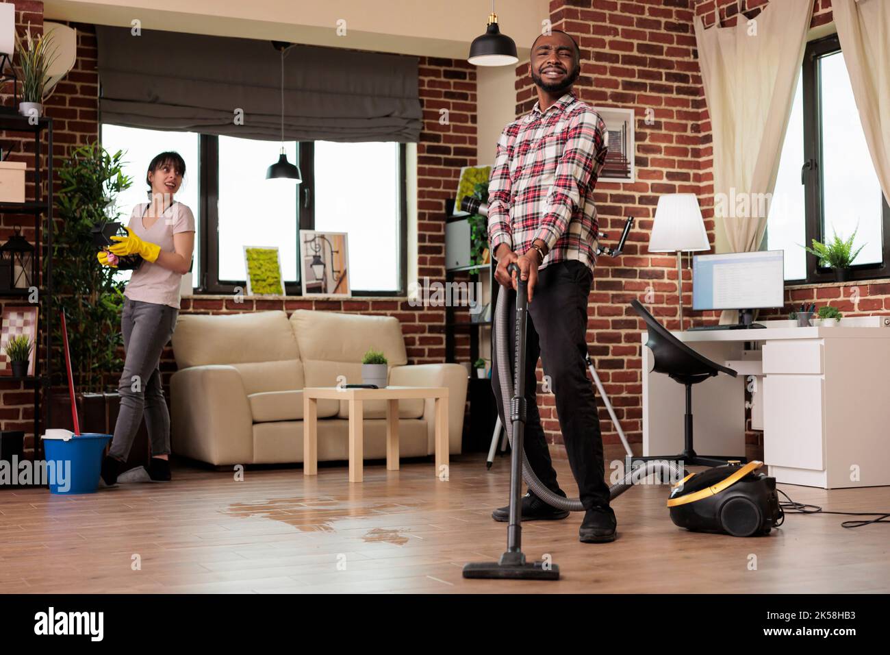 Multiracial couple happily cleaning at home, african american man vacuuming the whole place while woman dusts the shelves. Working as a team to keep house clean and tidy. Stock Photo