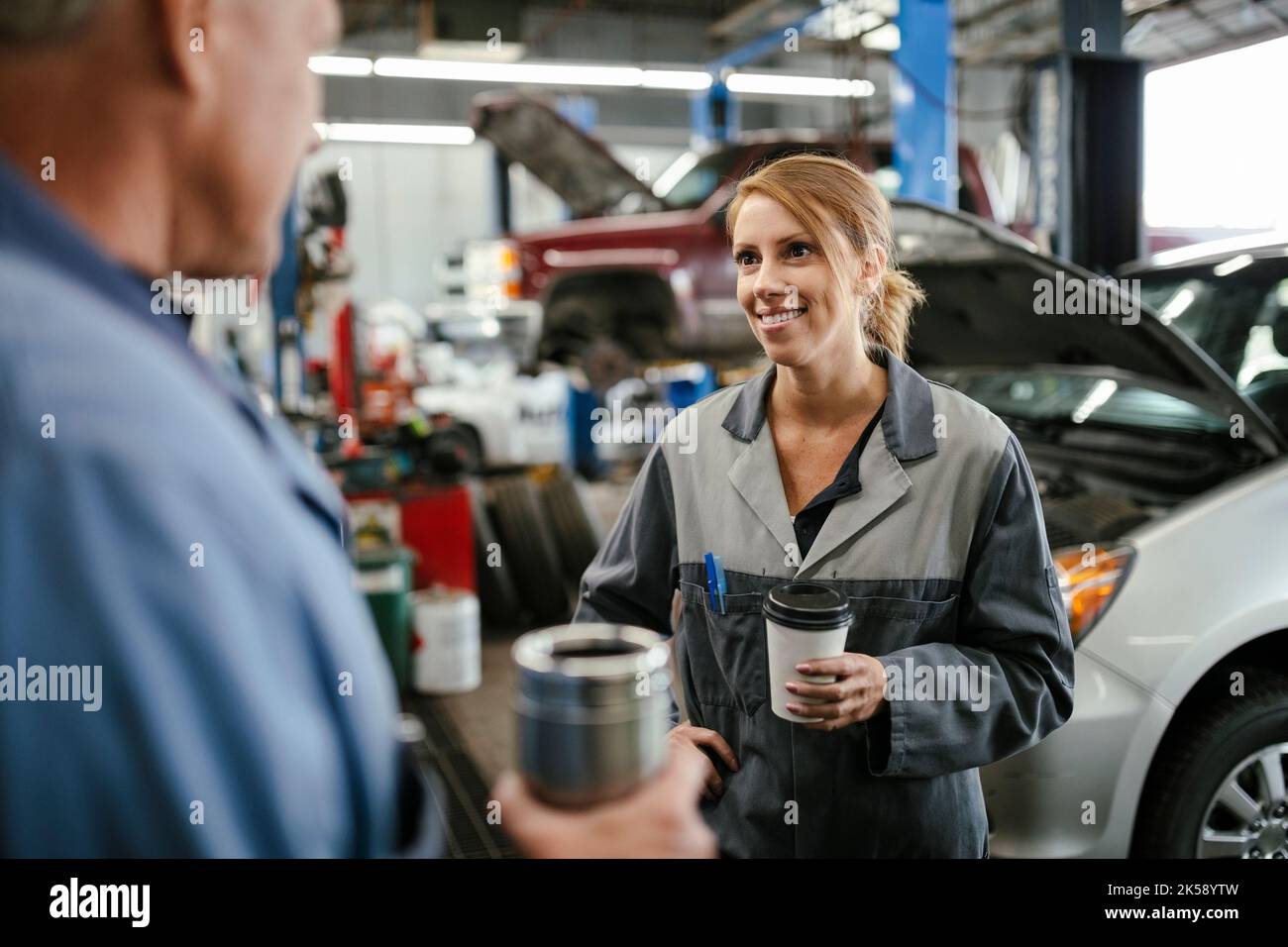 Female car mechanic taking coffee break in garage talking to colleague