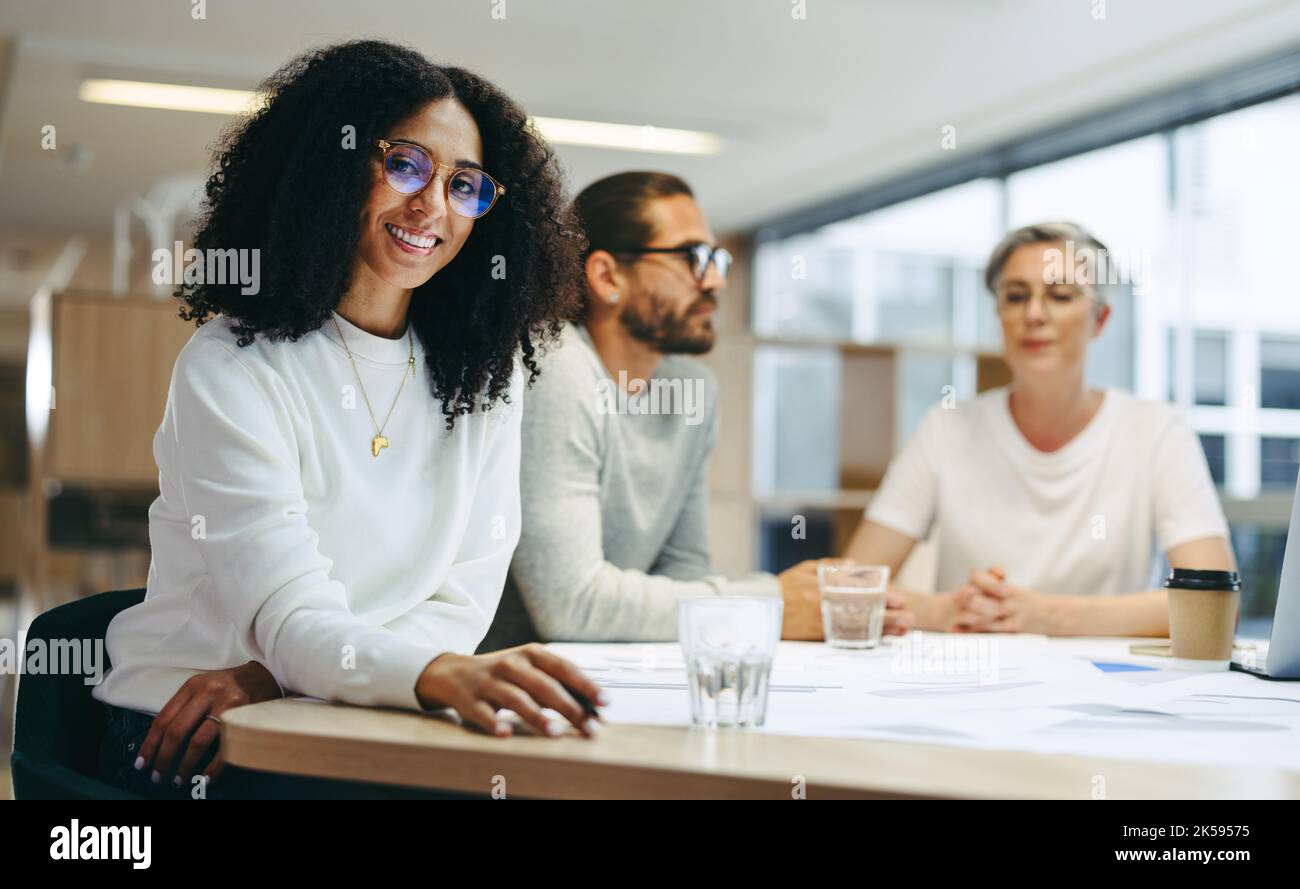 Cheerful young businesswoman smiling at the camera while sitting in a meeting with her team. Group of innovative design professionals sharing creative Stock Photo