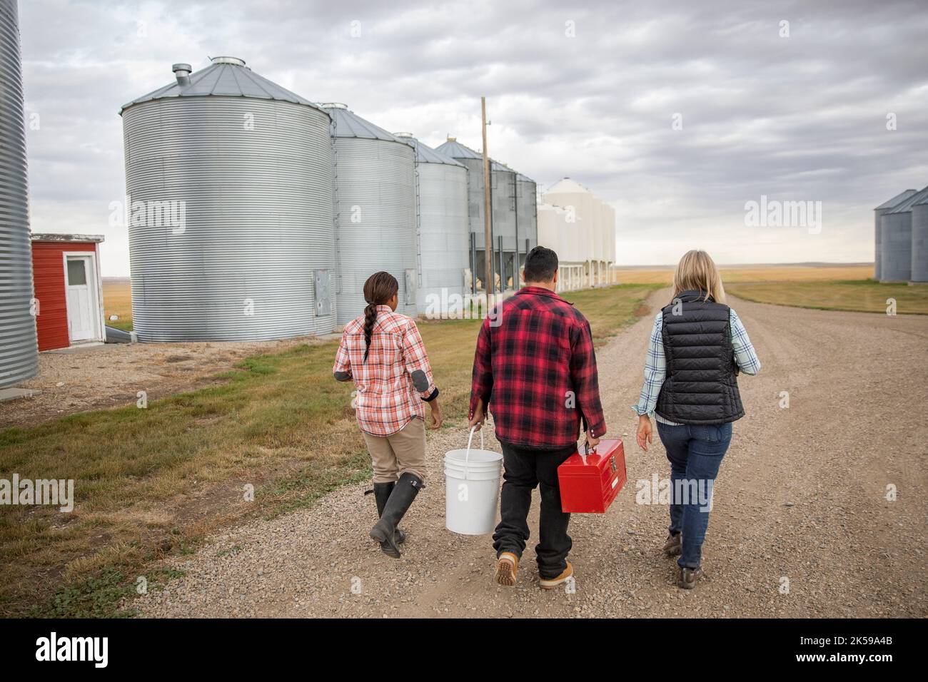 Farmers with toolbox and bucket walking along silos on gravel road on