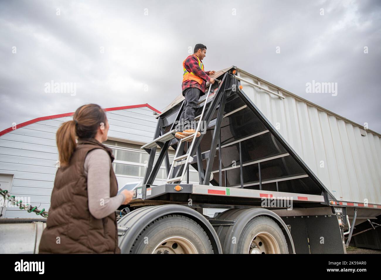 Farmer with digital tablet talking with farmworker on trailer ladder Stock Photo