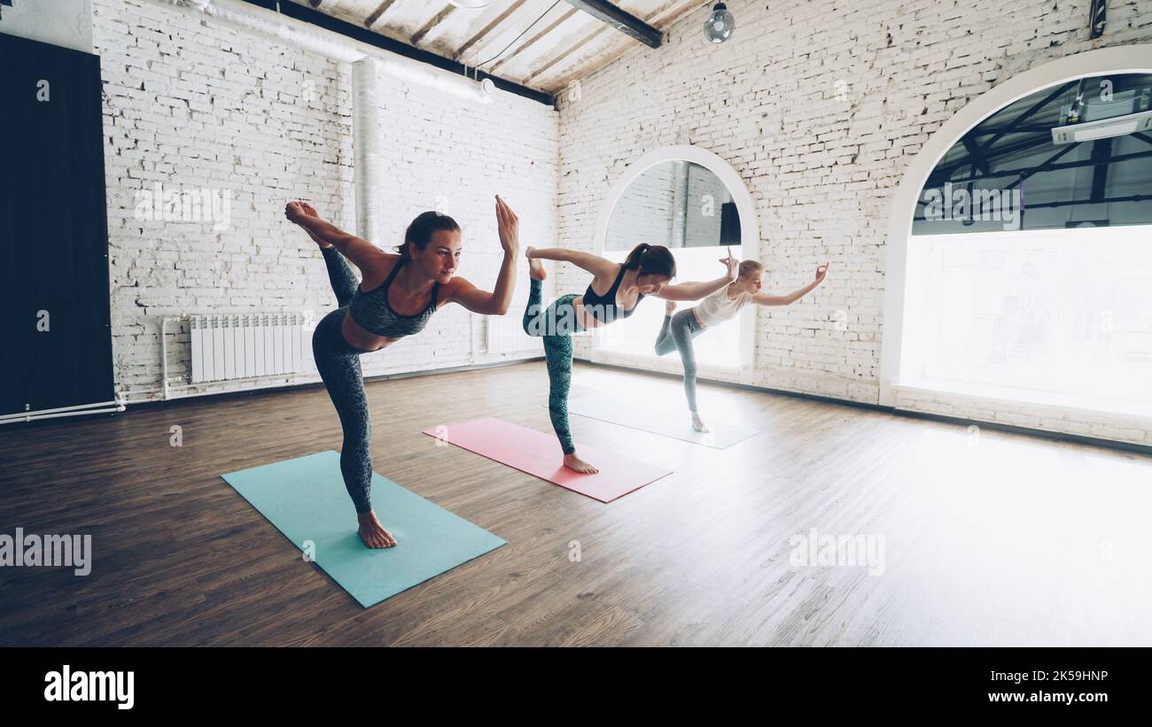 Dolly shot of pretty slim young women doing balancing yoga exercises in light loft style gym. Students are concentrated on practice. Sports equipment, fitness balls and mats are visible. Stock Photo