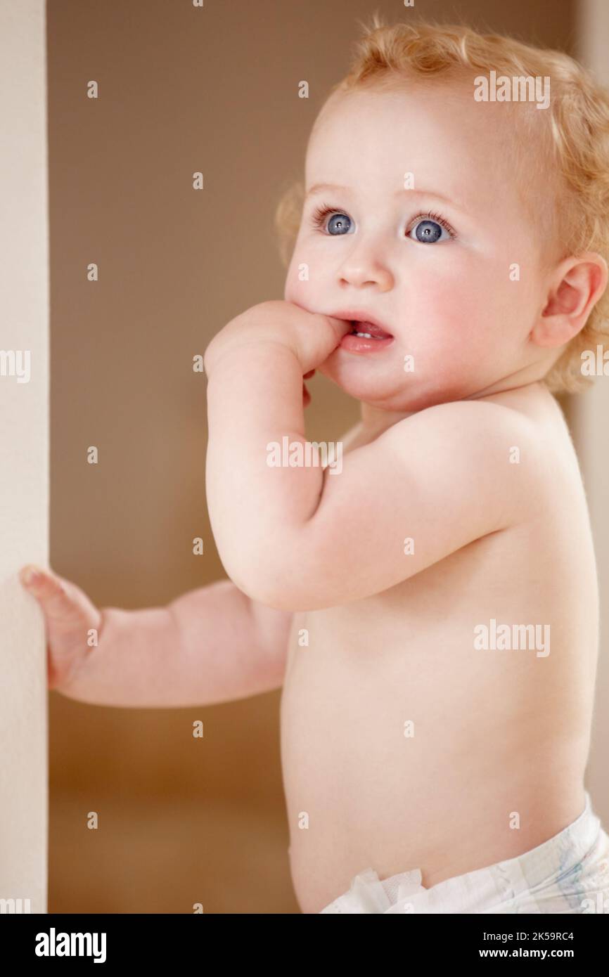 So curious about his world. Cute baby boy sucking on his hand while standing indoors. Stock Photo