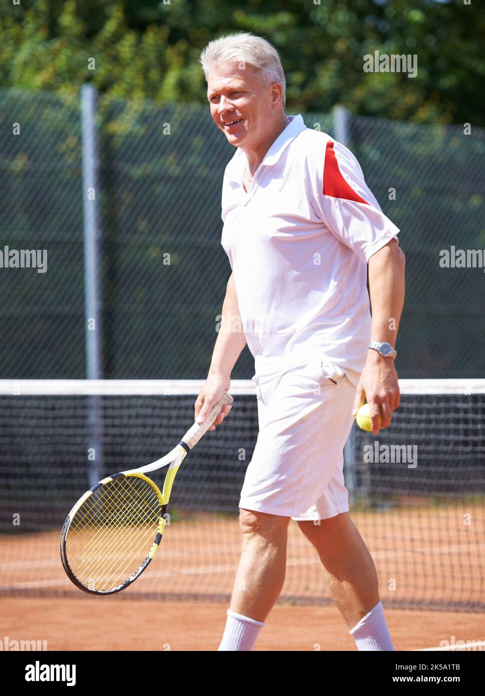 Tennis has always been his favorite past-time. A senior man walking on a clay court during a game of tennis. Stock Photo