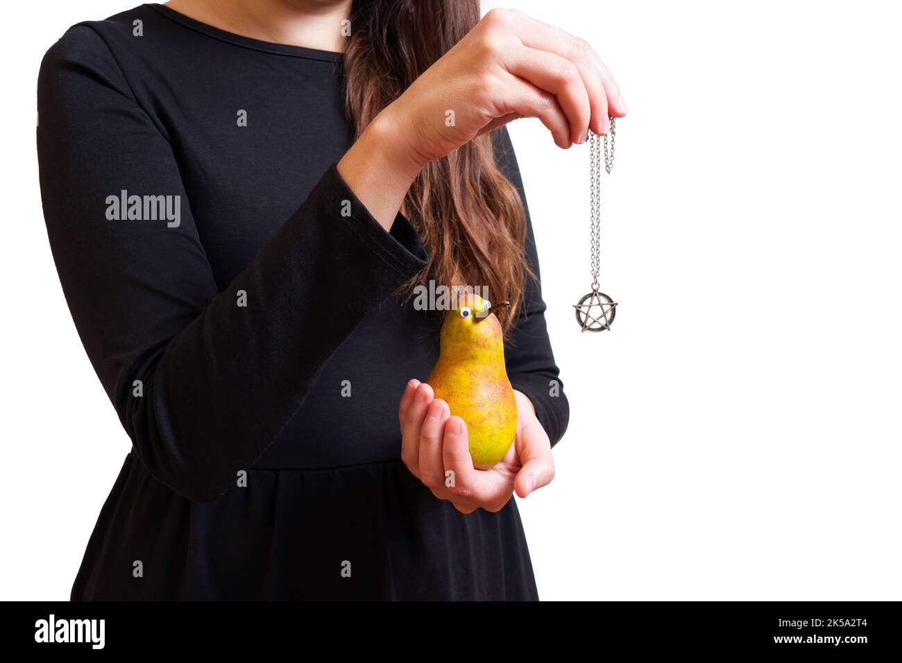 Young witch in black holds an anthropomorphic pear character and pentagram necklace in hands making a spell. Creative reincarnation ritual concept. Stock Photo