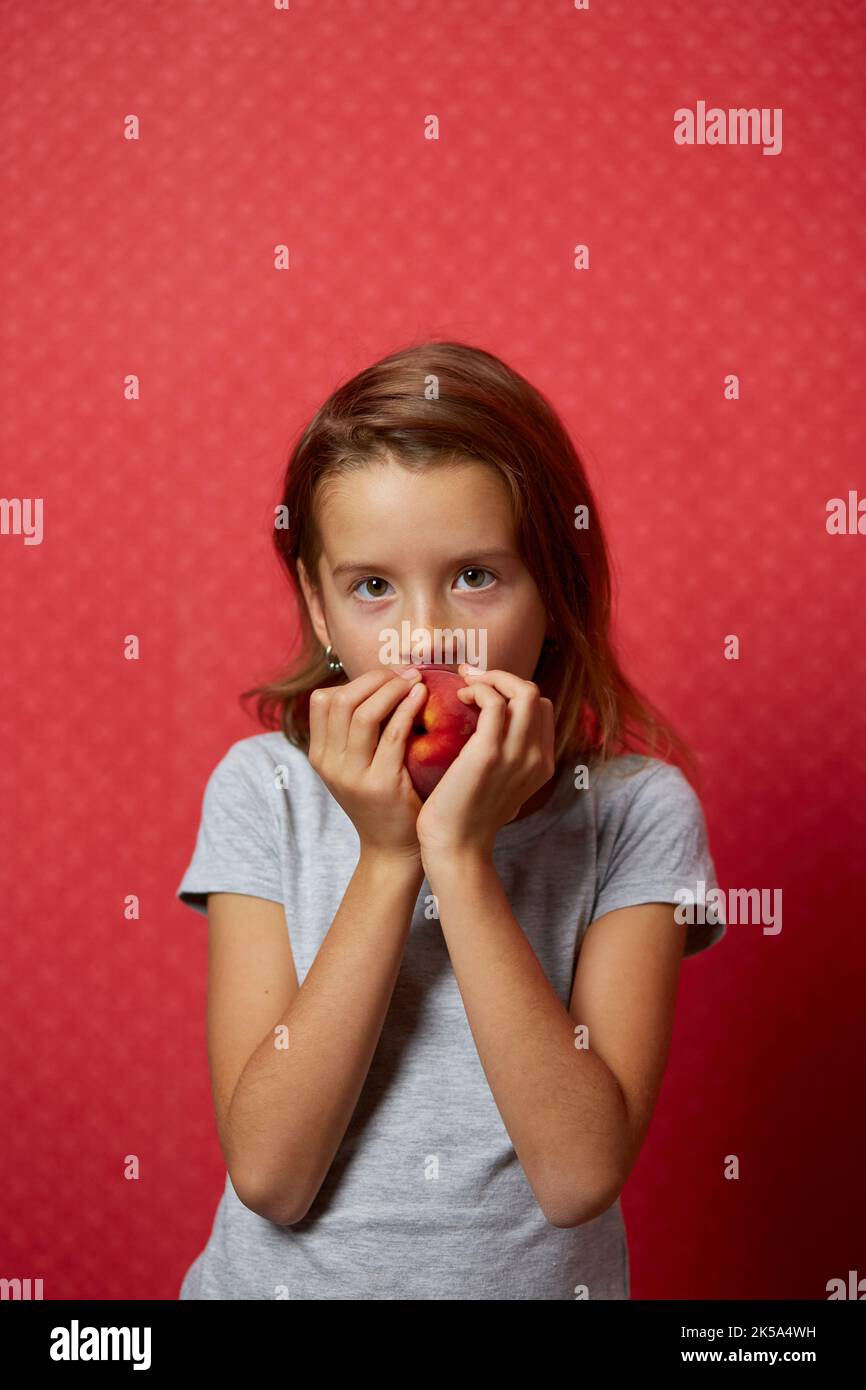 Portrait happy girl eats a peach on a red background, bites a juicy