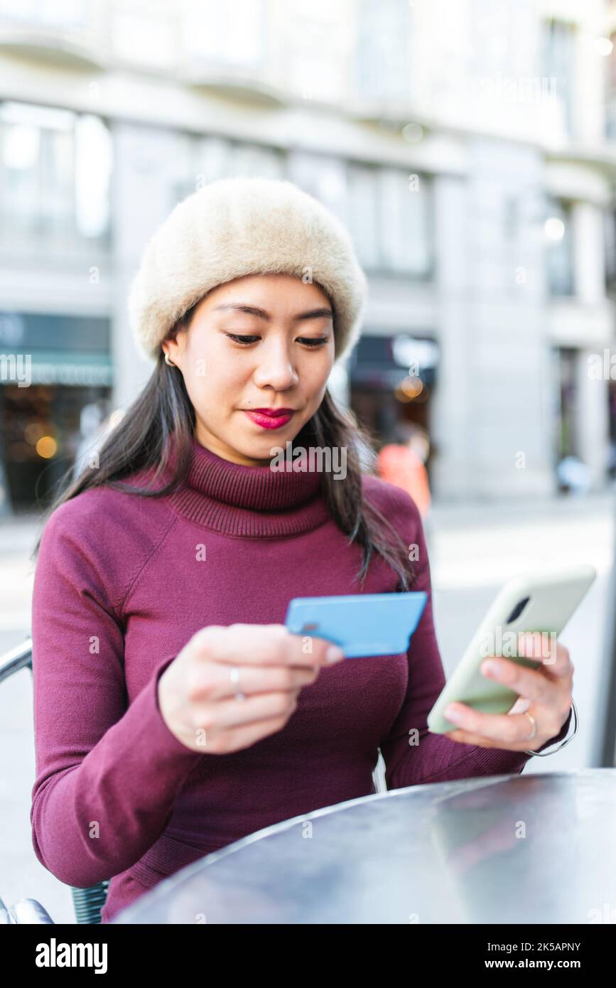 Content Asian female with credit card making online purchase via smartphone while sitting on terrace of outdoor cafe in city Stock Photo