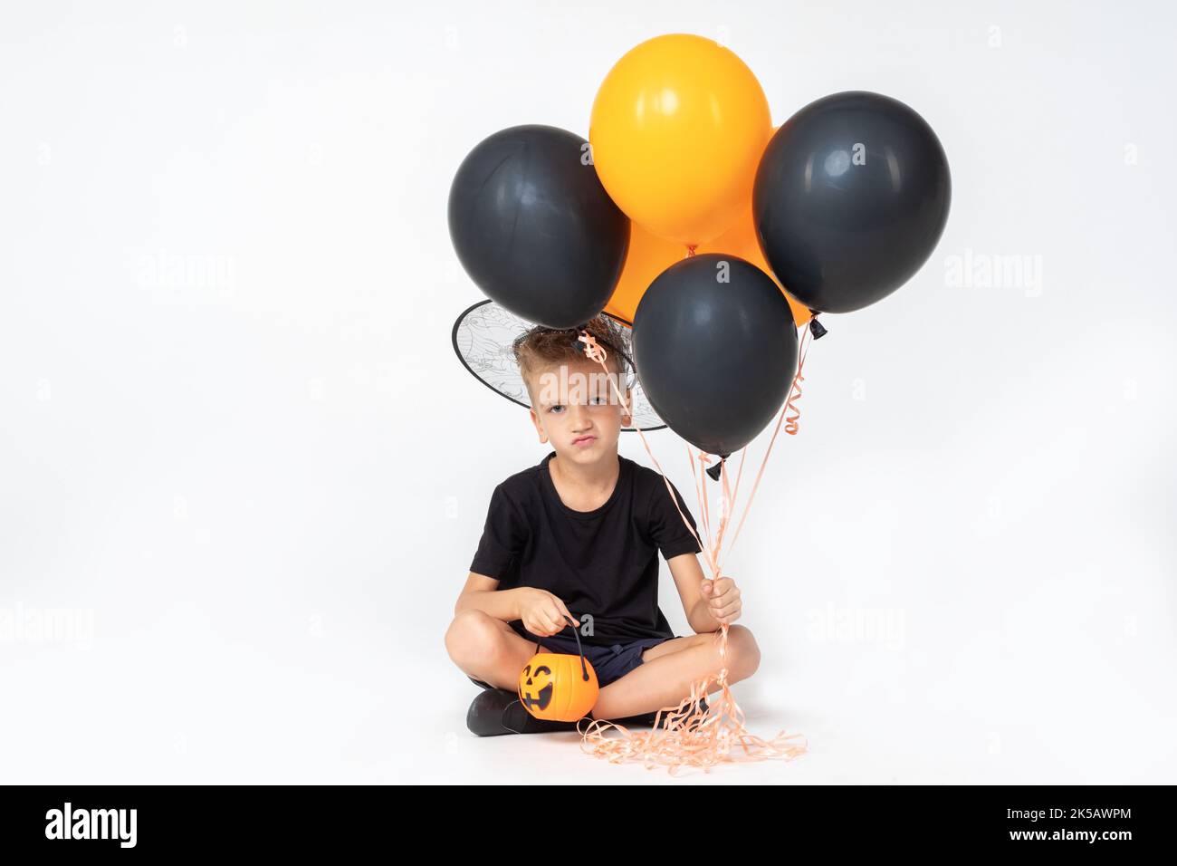 A cheerful happy boy in a wizard's hat holding a pumpkin-shaped bucket and black and orange balloons, sitting on a white studio background. Ready for Stock Photo