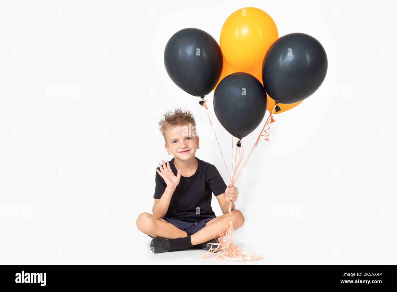 A cheerful happy boy in a wizard's hat greets a waving hand holding black and orange balloons, sitting on a white studio background. Hello Halloween Stock Photo