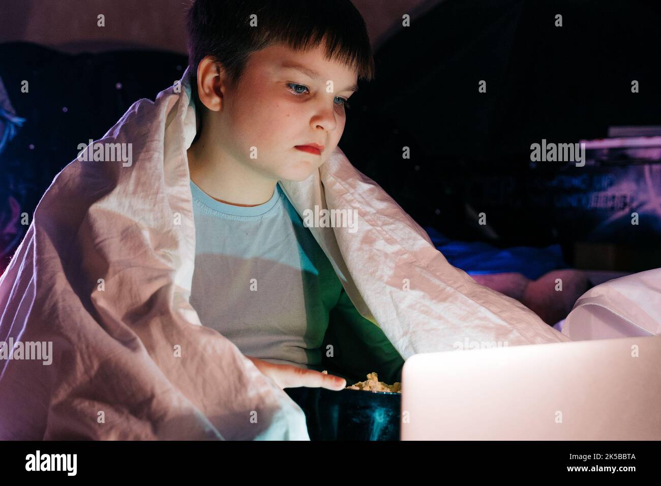 Happy kid son laughing eat popcorn remote control watching funny comedy tv show sitting on sofa having fun viewing video on laptop in evening at home. Stock Photo