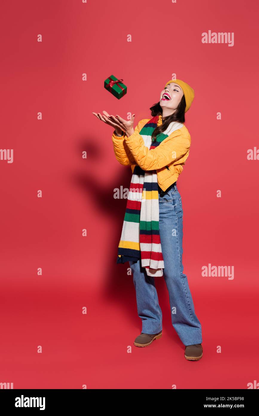 full length of amazed woman in beanie hat and scarf catching wrapped gift box on red,stock image Stock Photo
