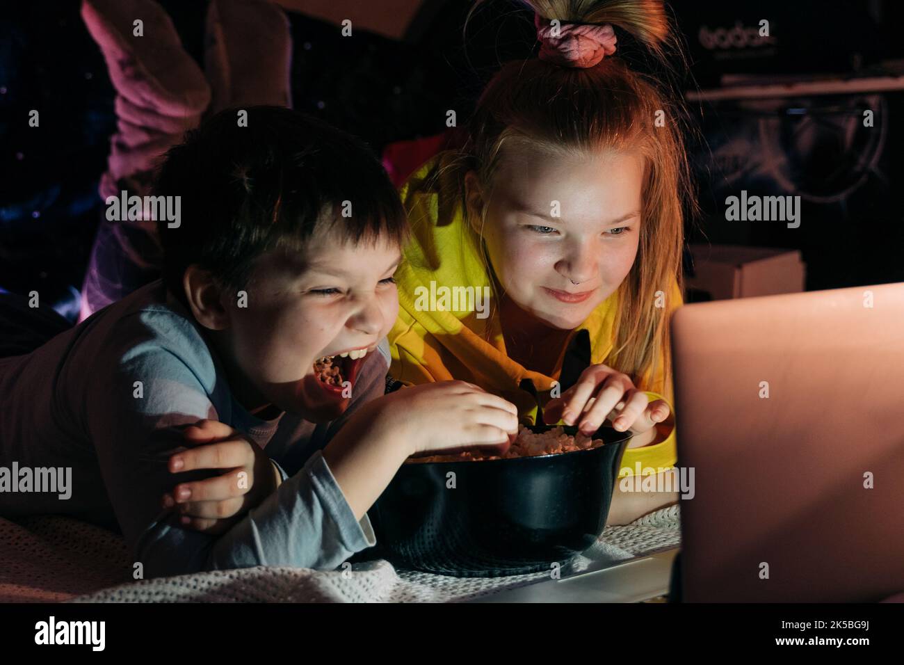 Overjoyed Kids sister brother laughing spend weekend free time together at home on couch bed eating popcorn. teen children watching video cartoon use Stock Photo