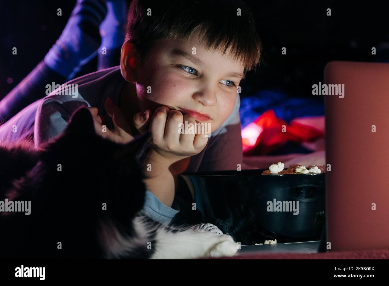 Cheerful teen boy using laptop at home watching funny movie cartoon eat pop corn, browsing internet on sofa in living room. Smiling kid looking at scr Stock Photo