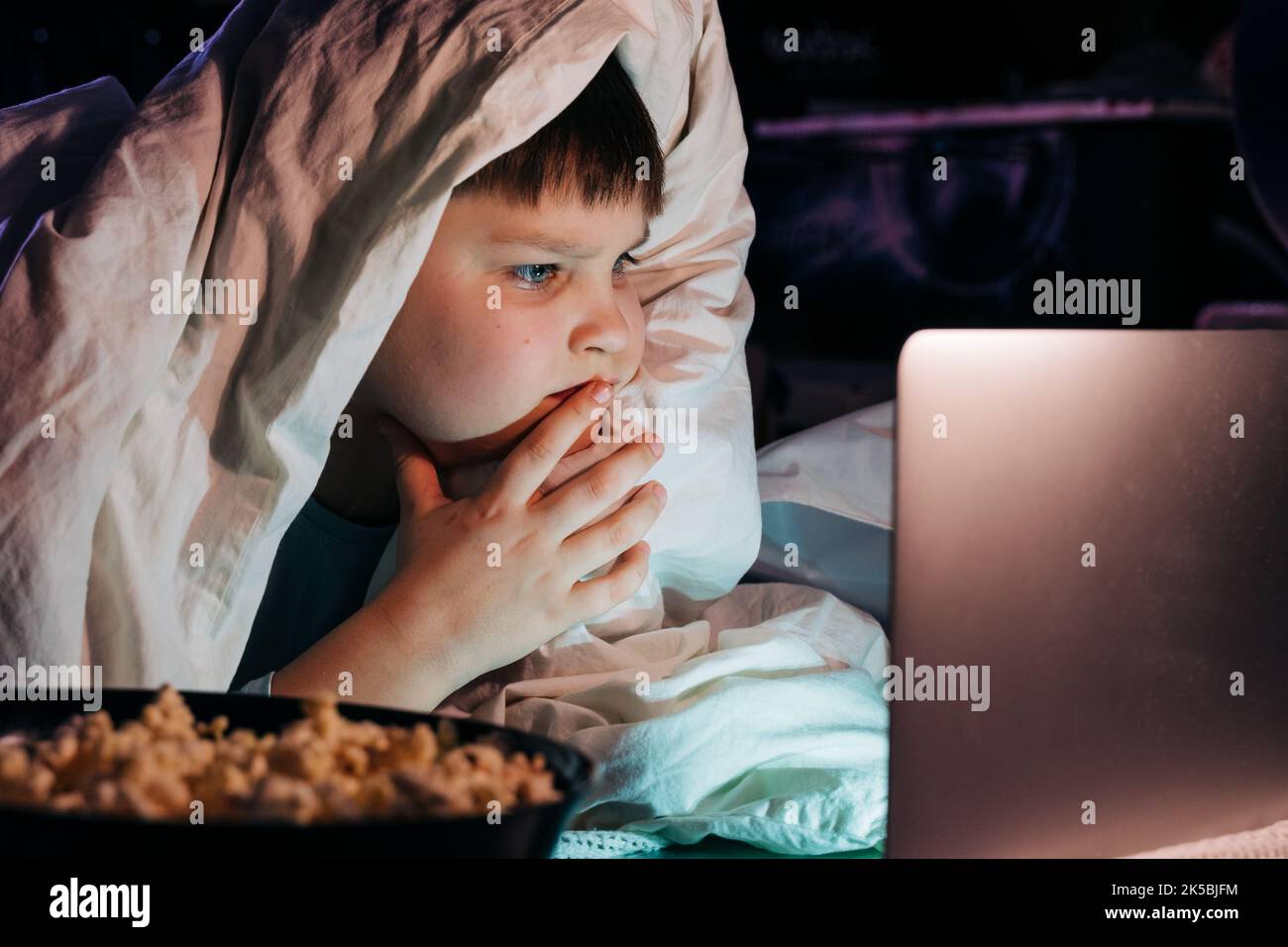 Happy kid son laughing eat popcorn remote control watching funny comedy tv show sitting on sofa having fun viewing video on laptop in evening at home. Stock Photo