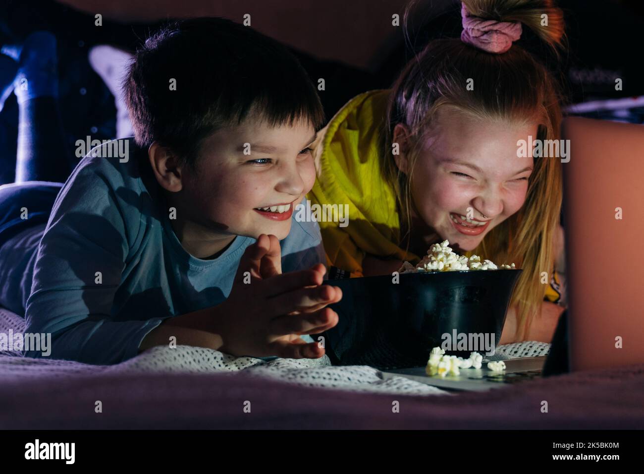Overjoyed Kids sister brother laughing spend weekend free time together at home on couch bed eating popcorn. teen children watching video cartoon use Stock Photo