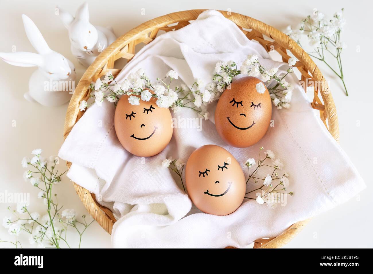 Three happy Easter eggs with cute faces in floral wreath crowns on white background in nest and two white bunnies. Easter eggs with flowers and sleepy Stock Photo