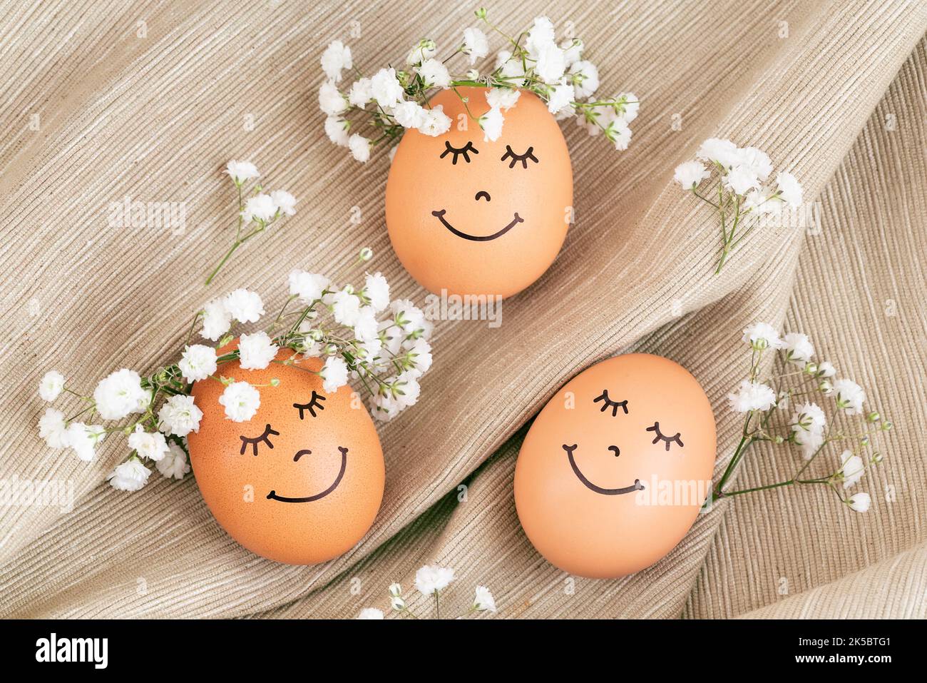 Three happy Easter eggs with cute faces in floral wreath crowns on brown burlap background. Easter eggs with flowers and sleepy eyes in sunny light. H Stock Photo