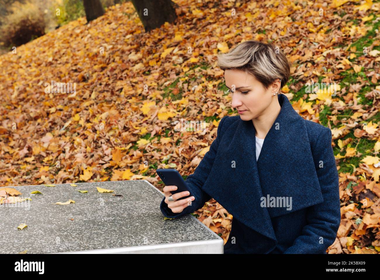 Short-haired blonde uses a smartphone sitting at a table in the park in the fall. Stock Photo