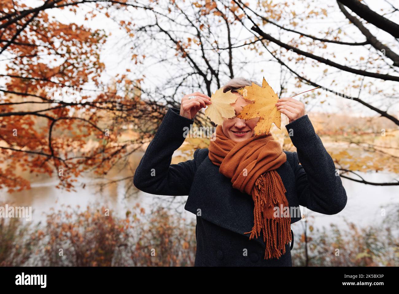 A woman in a coat and scarf in an autumn park covers her face with maple leaves. Stock Photo