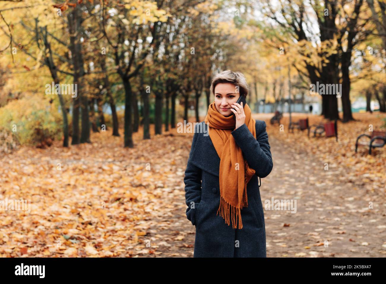 Short-haired blonde communicates on the phone walking in the autumn park in a coat and scarf. Stock Photo