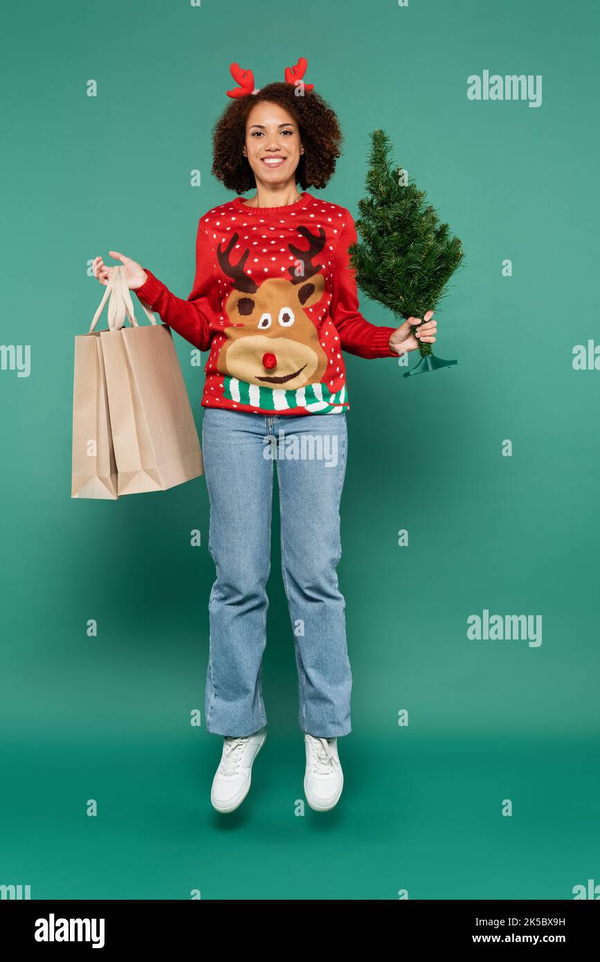 african american woman in festive outfit levitating with shopping bags and small christmas tree on green background,stock image Stock Photo
