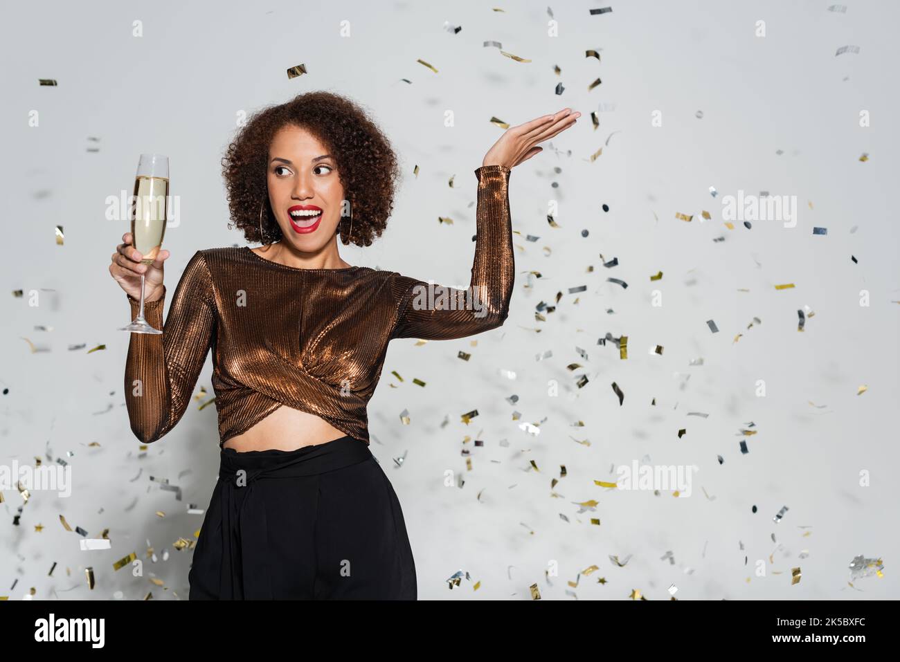 excited african american woman in festive clothes posing with champagne glass near confetti on grey background,stock image Stock Photo