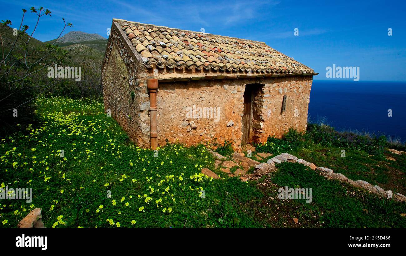 Italy, Sicily, Zingaro National Park, spring, green meadow, yellow flowers, stone hut with tiled roof, blue sea, blue sky Stock Photo