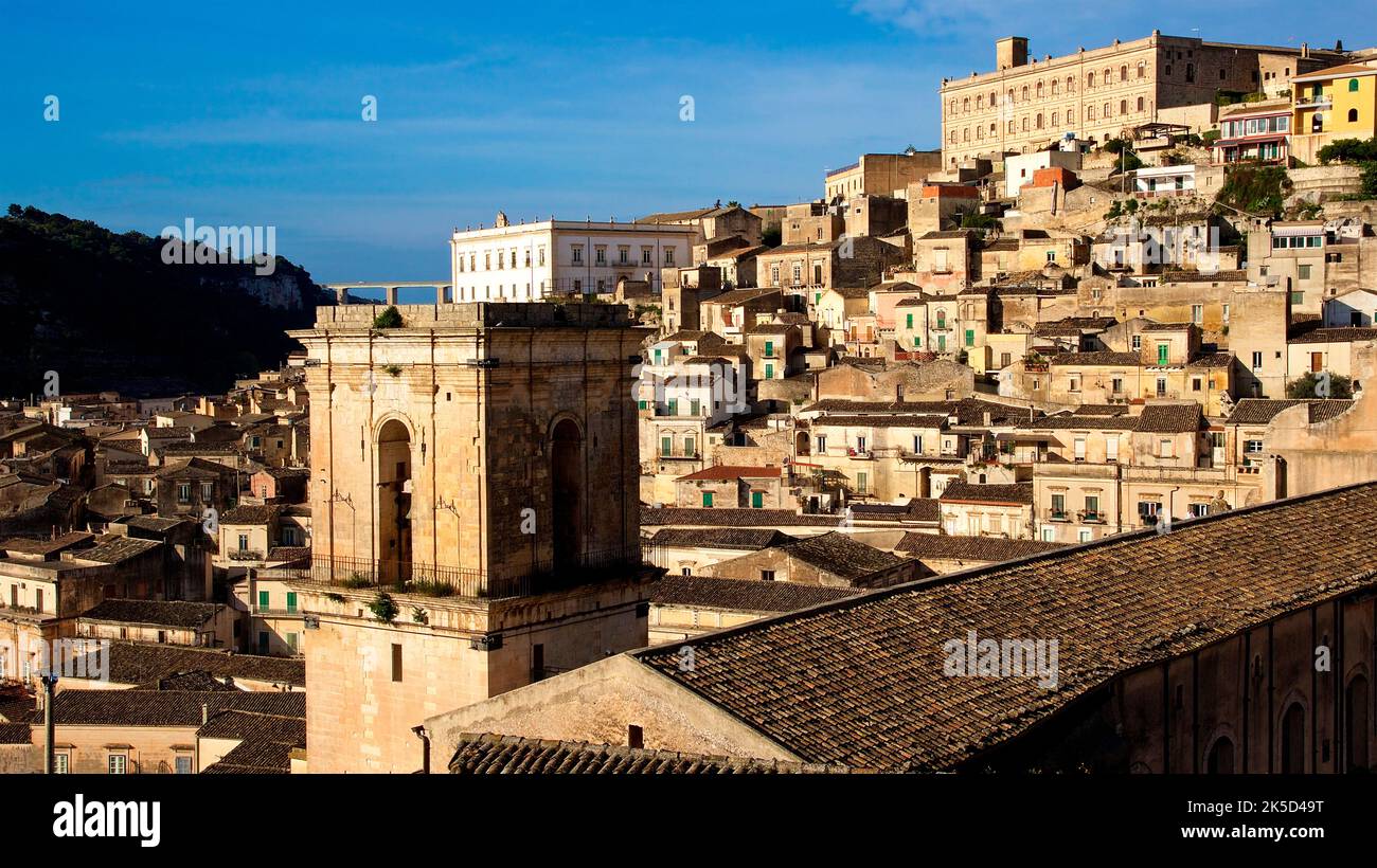 Italy, Sicily, baroque angle, baroque town, Modica, old town, evening light, rectangular tower in foreground, red tiled roof, buildings of old town behind, blue sky with veil clouds Stock Photo