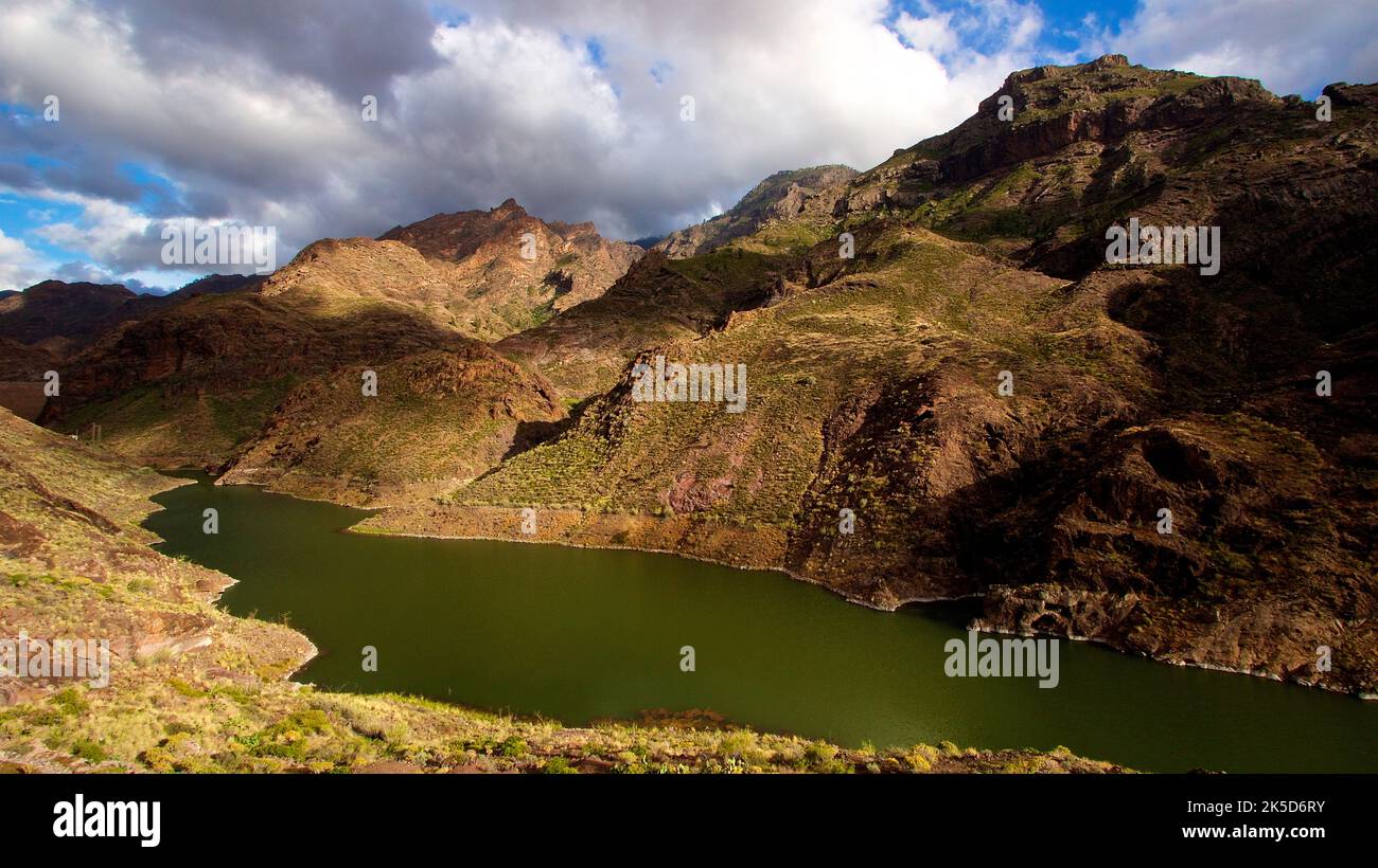 Spain, Canary Islands, Gran Canaria, Barranco de la Aldea, reservoir press of Parralilo, viewpoint Mirador del Molina, dark green water surface, play of light and shadow on the canyon walls, slopes green overgrown, blue sky with gray-white clouds Stock Photo