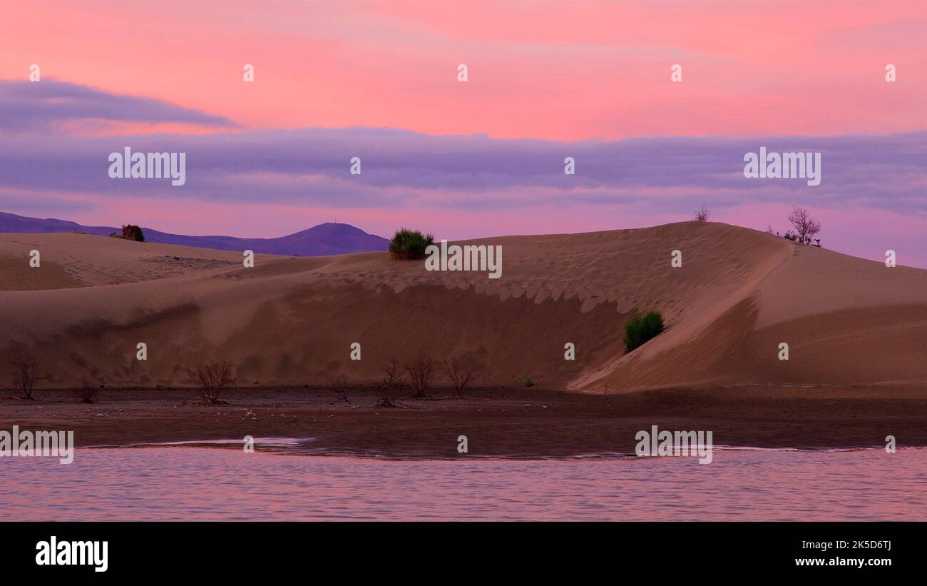Spain, Canary Islands, Gran Canaria, south coast, Maspalomas, dune area, dusk, pink sky, dunes in the middle ground, mountain peaks in the background, shallow sea water in the foreground Stock Photo