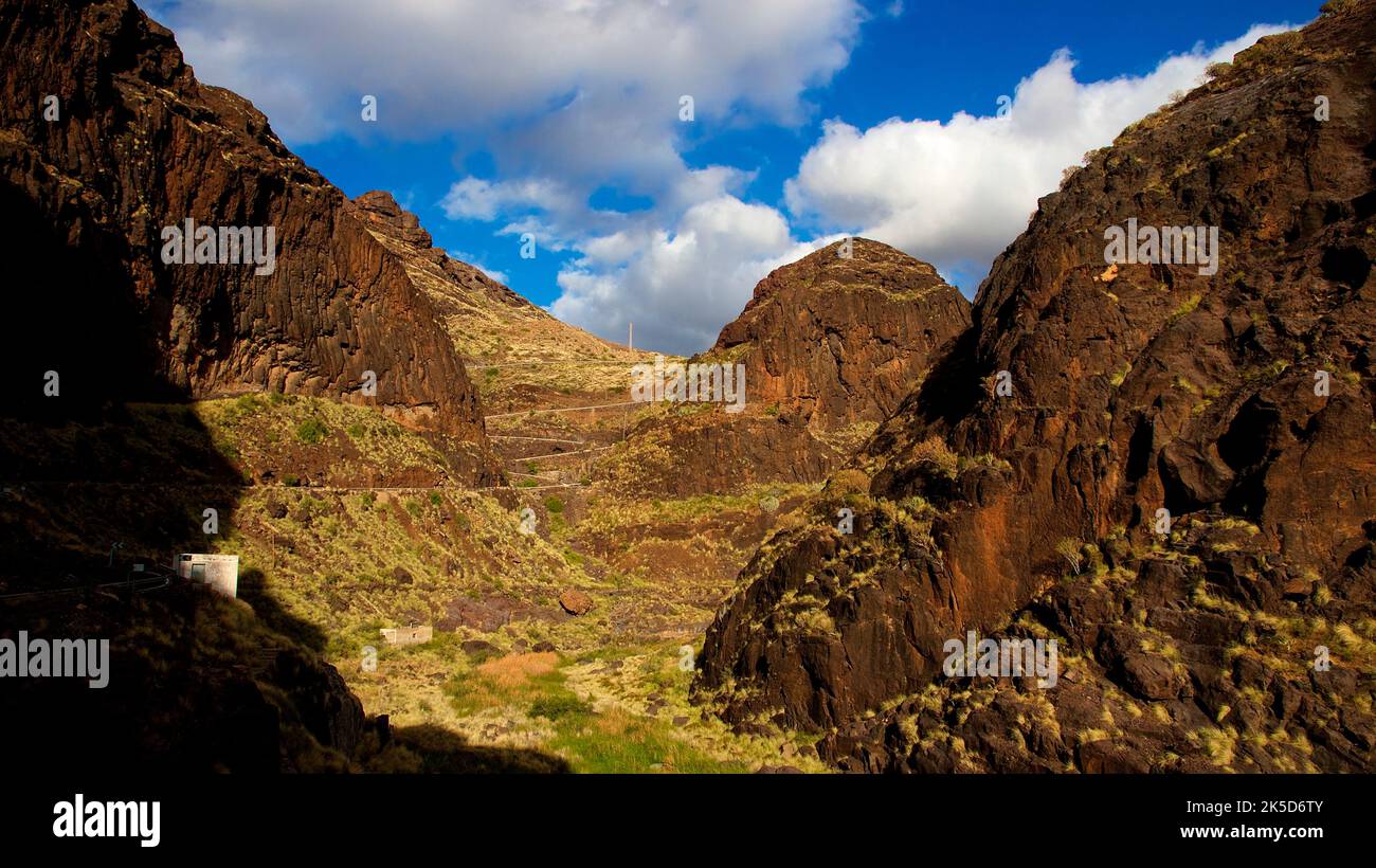 Spain, Canary Islands, Gran Canaria, Barranco de la Aldea, view up north into the gorge, winding narrow road winding down a steep slope, overgrown slopes, blue sky with white clouds Stock Photo
