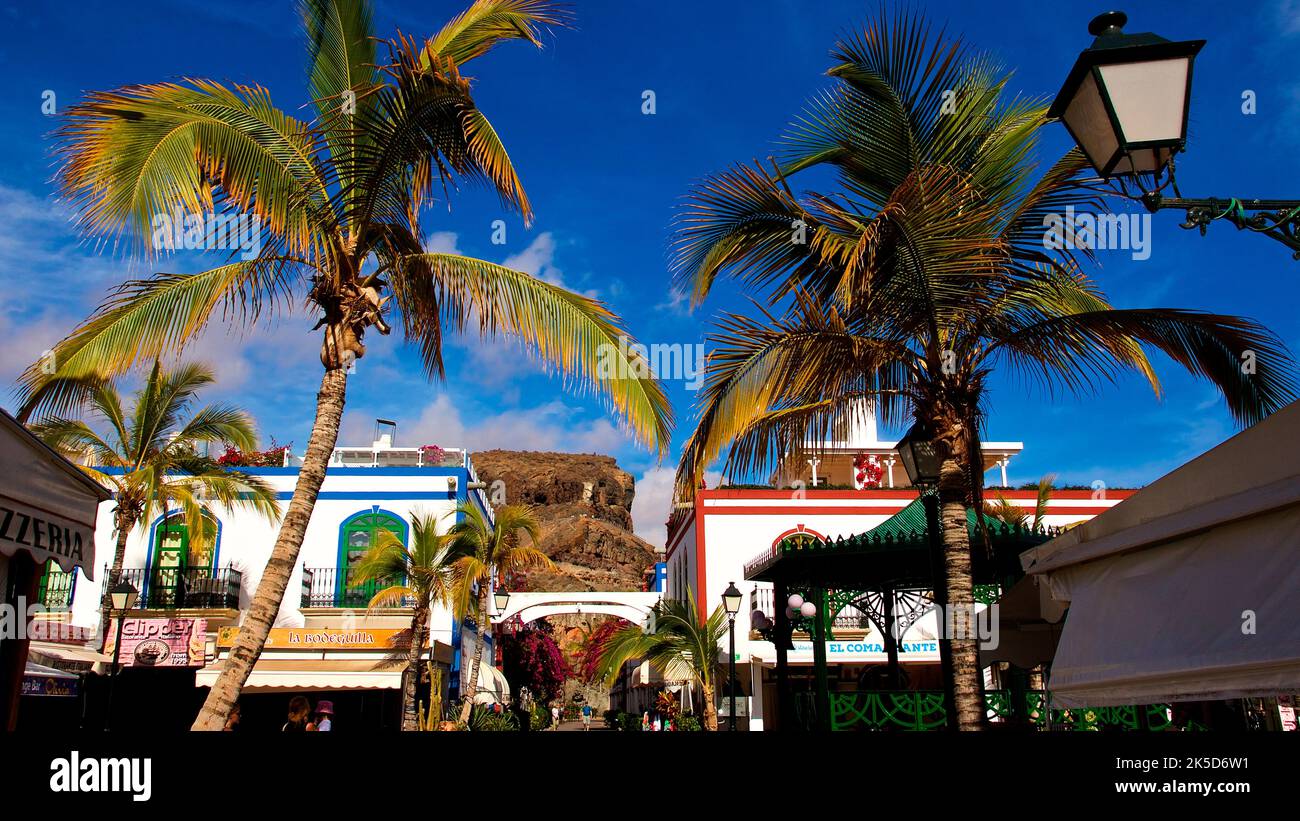 Spain, Canary Islands, Gran Canaria, southwest coast, Puerto de Morgan, Little Venice, wide angle shot, palm trees and lantern in foreground, white buildings with colored ornamental stripes in middle ground, hills ocher in background, dark blue sky, few white clouds Stock Photo