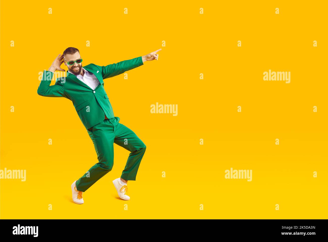 Happy funny man in green suit pointing at copy space while dancing on yellow background Stock Photo
