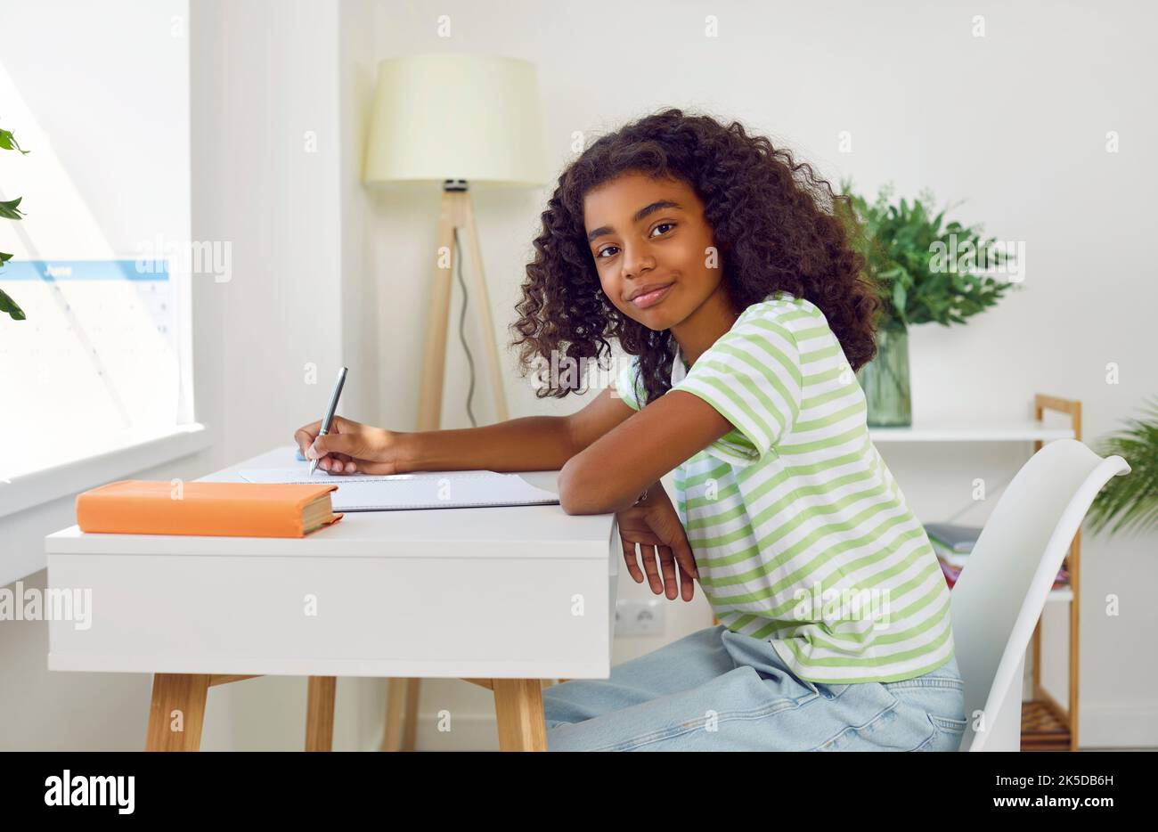 Positive teen african american girl is doing her homework sitting at desk at home lookinh at camera. Stock Photo