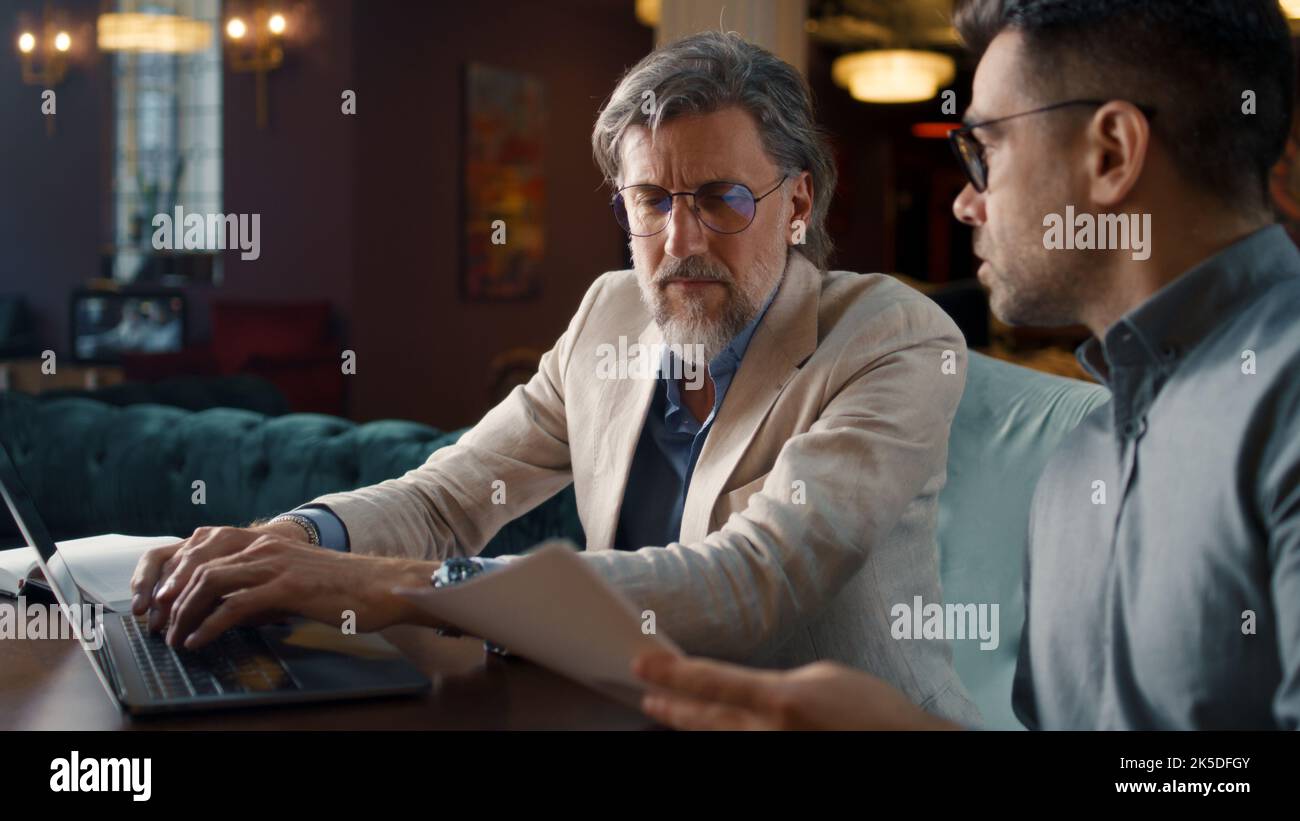 Male colleagues talking, using laptop, working with papers and documents, having business meeting in office restaurant. Teamwork on strategy of E-commerce company Stock Photo