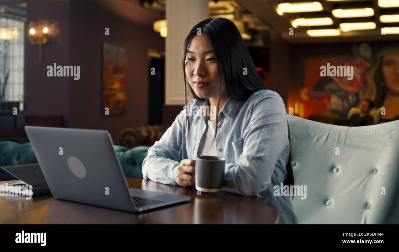 Asian businesswoman looking at screen of laptop, watching business training, working online on startup project, drinking coffee in office cafe. Worker of E-commerce marketing company Stock Photo