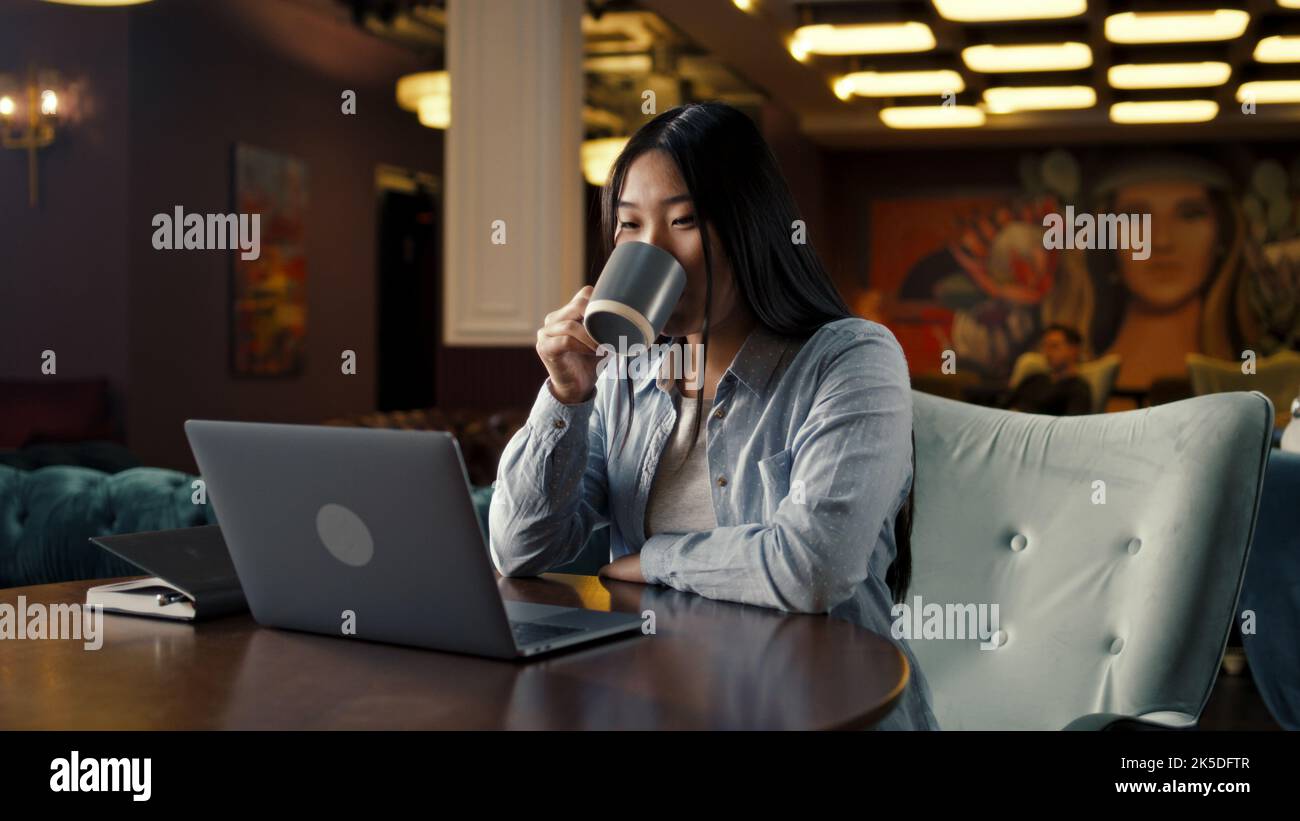 Asian businesswoman looking at screen of laptop, watching business training, working online on startup project, drinking coffee in office cafe. Worker of E-commerce marketing company Stock Photo