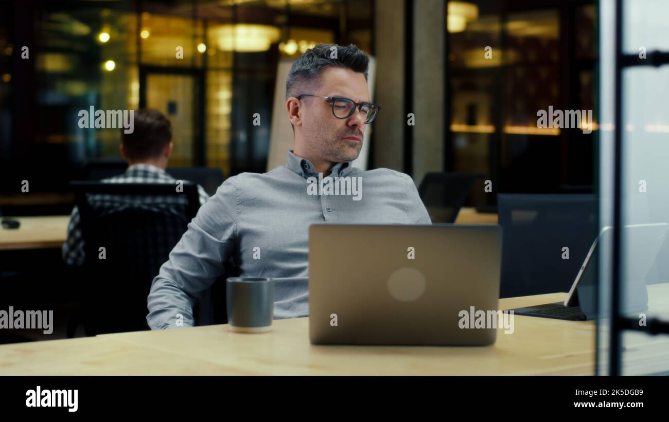 Creative male office worker listening to music, using laptop and digital tablet computer, working in modern office near colleagues on startup project. Work on business strategy of company Stock Photo