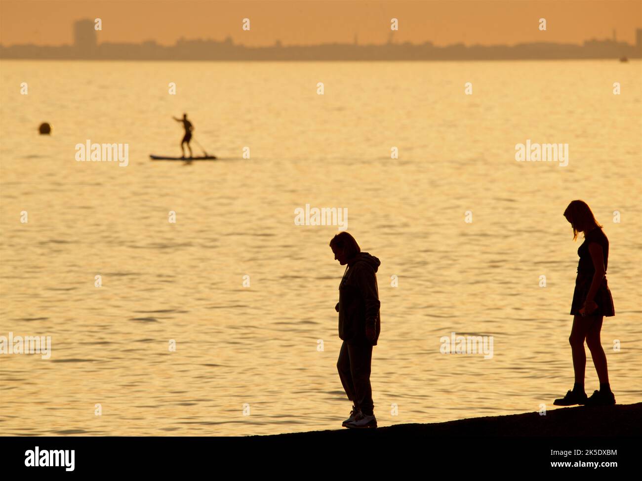Silhouetted shapes of people standing on a groyne on a beach, Brighton & Hove. The sky aglow with the fading light of the day. Paddleboarders on the sea in the distance. England, UK Stock Photo