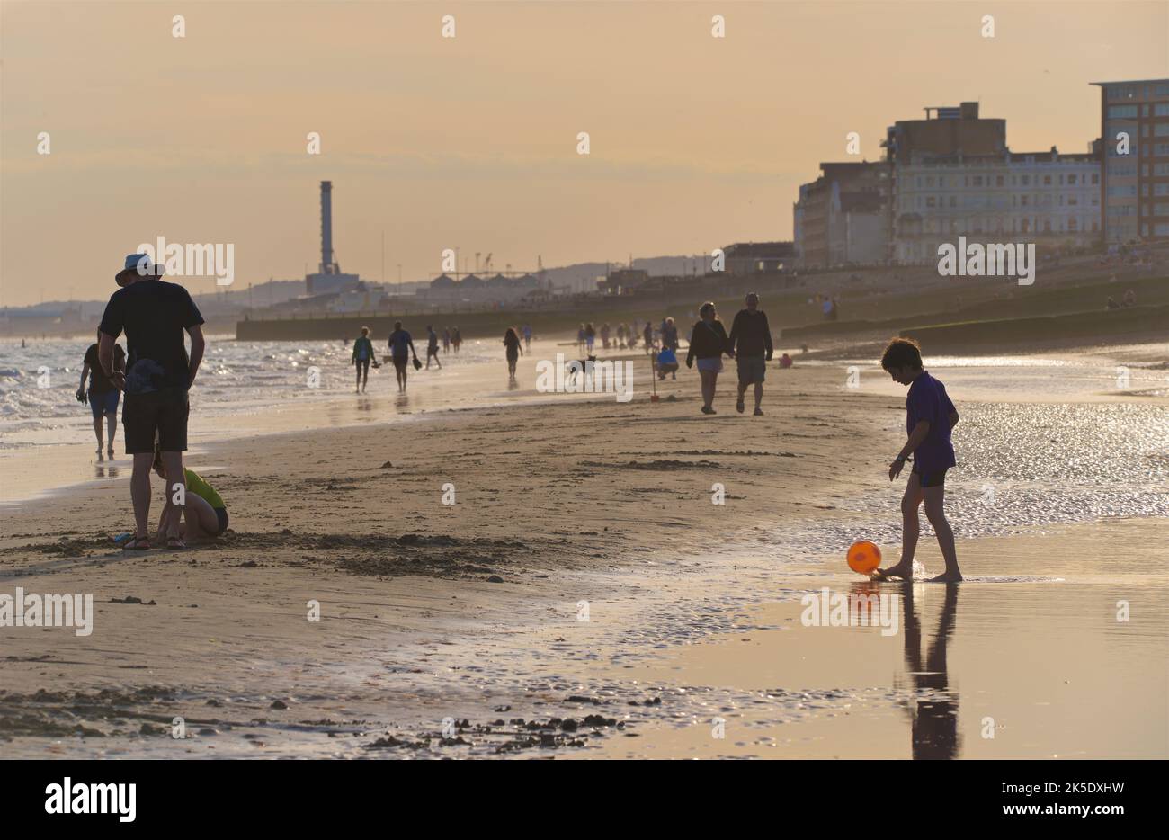 Silhouetted shapes of people on the beach at low tide in the late afternoon, July, Brighton & Hove, East Sussex, England, UK. Child with orange bootball Stock Photo