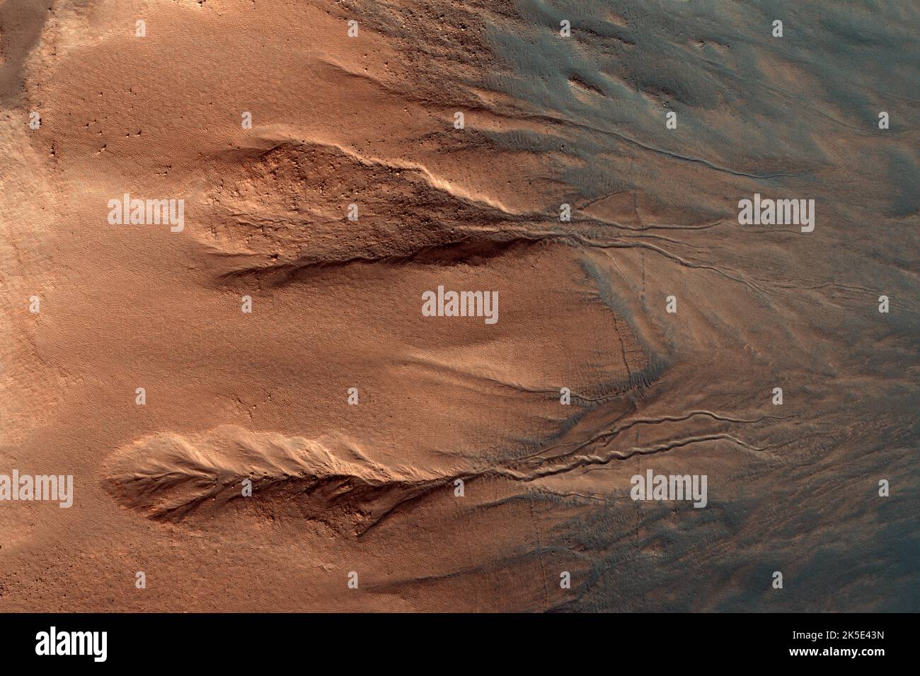MARS. Gullies are relatively common features in the steep slopes of crater walls, possibly formed by dry mass movement, movement of carbon dioxide frost, or perhaps the melting of ground ice. This example shows a section of crater wall from the rocky crater rim at the far left of the image, down to the dark dusty dunes on the crater floor in the bottom right. The rock of the crater walls shows up deep orange, and the sandy deposits on the crater floor and the base of the crater walls appear blue (not actual colour.)  An optimised version of NASA imagery. Credit: NASA/JPL/UArizona Stock Photo