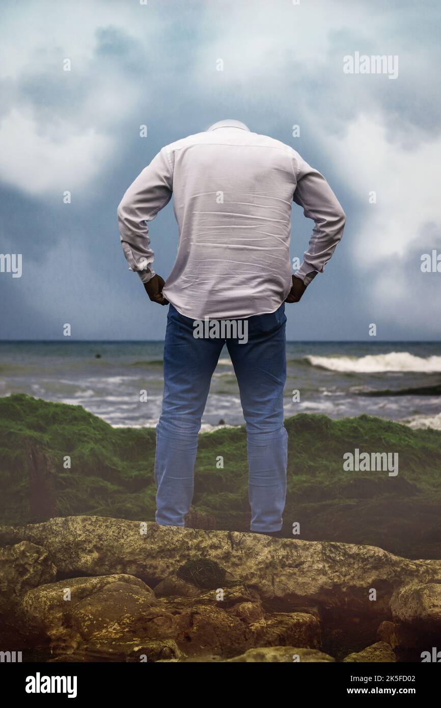 a headless man in a white shirt and blue jeans standing on a coast facing a sea. Stock Photo